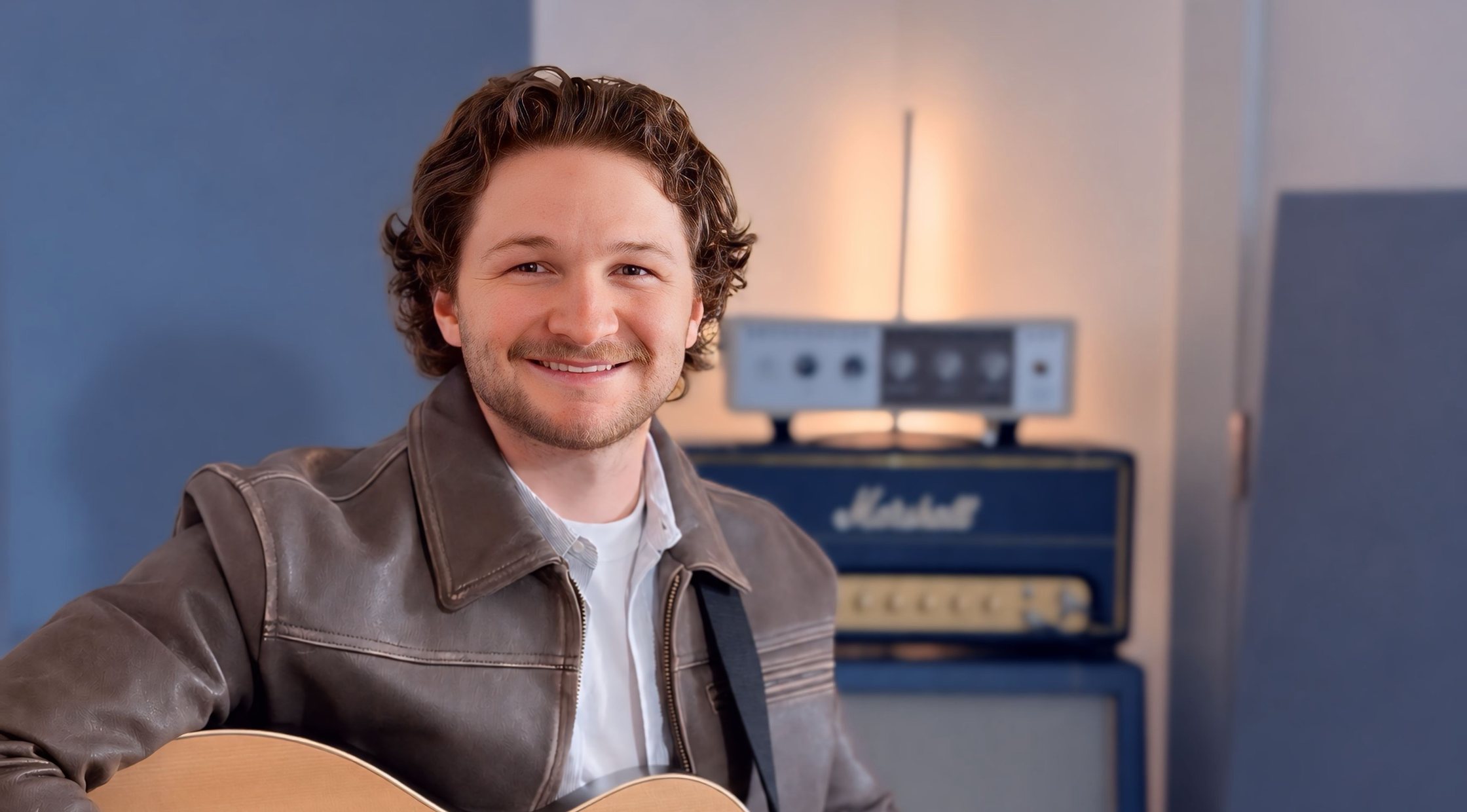 Corbin smiling and holding an acoustic guitar in his studio