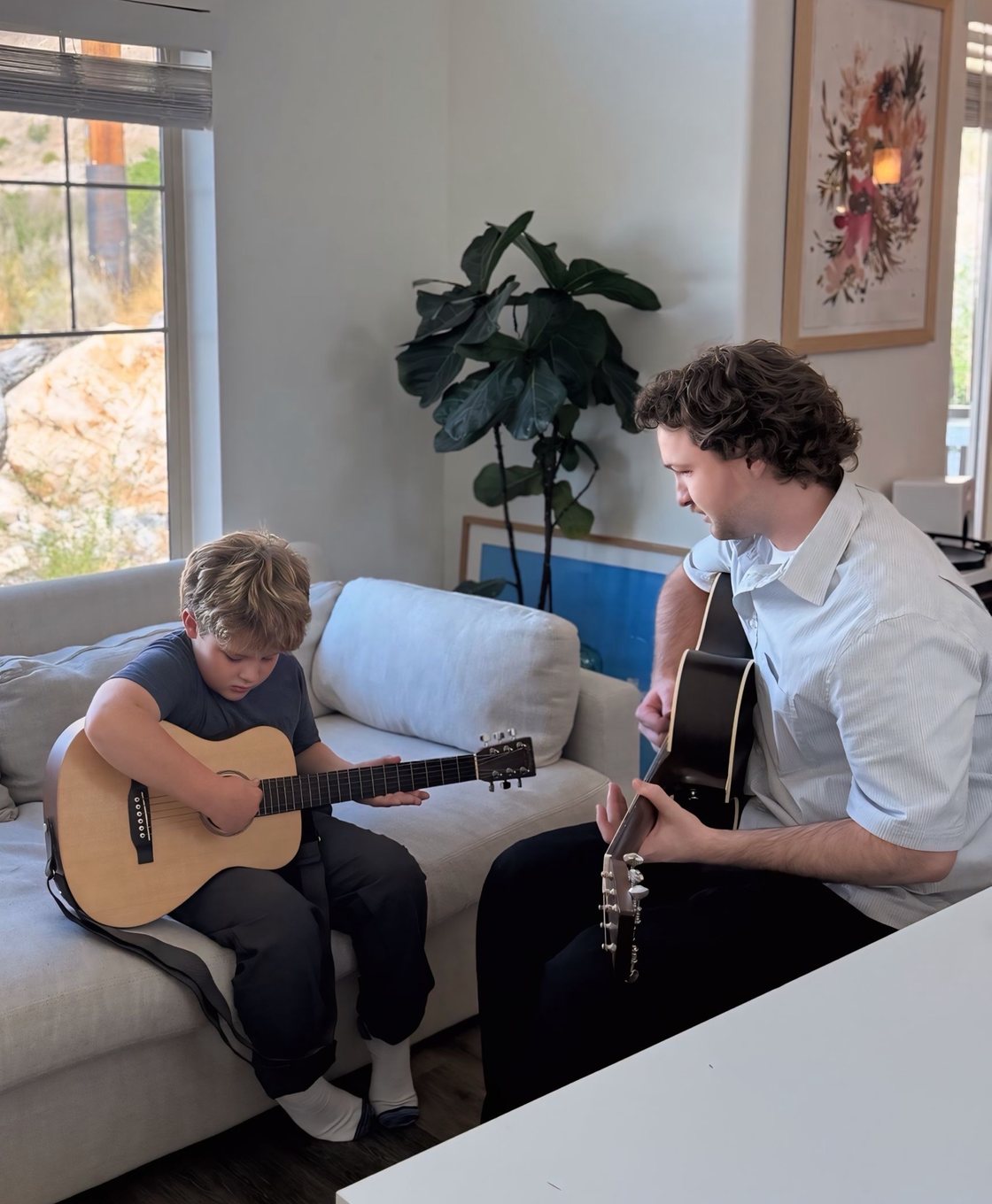 Corbin teaching a young student on the couch in the student’s home, both holding acoustic guitars